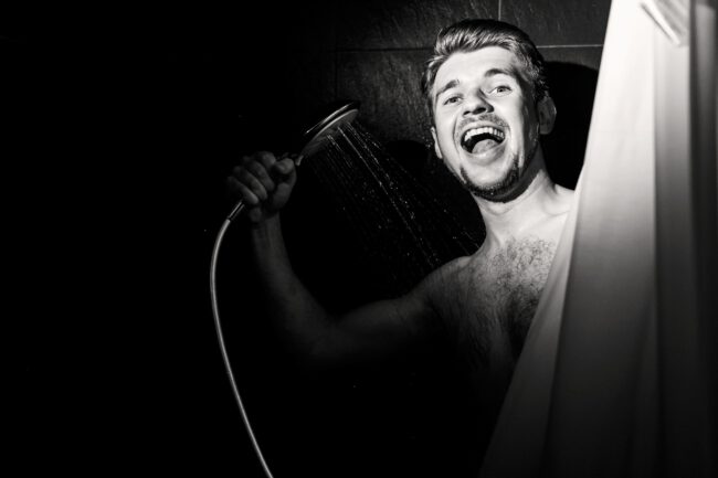 Man looks funny holding shower over him in black bathroom